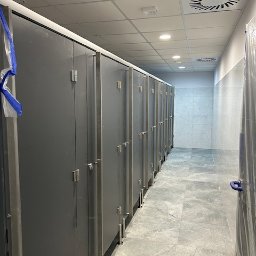 Budmar Marcin Garncarek - Rząd szarych kabin sanitarnych z metalowymi uchwytami w nowocześnie wykończonej toalecie publicznej, widok z perspektywy korytarza, podłoga wyłożona płytkami imitującymi kamień.