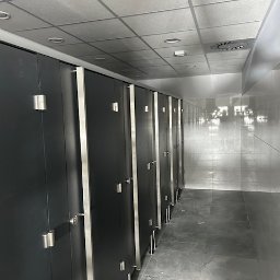 Budmar Marcin Garncarek - Rząd kabin toaletowych z szarymi drzwiami i metalowymi okuciami, widok wzdłuż długiego korytarza z szarą podłogą i sufitem kasetonowym.