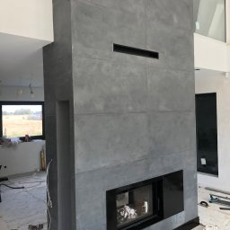 Budmar Marcin Garncarek - Wysoki kominek z szarych płyt imitujących beton, z czarnym wkładem kominkowym i czarnymi wstawkami, w surowym wnętrzu w trakcie prac wykończeniowych.
