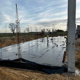 Budmar Marcin Garncarek - Wylewanie fundamentów pod budynek z widocznymi zbrojeniami, pracownikami w odblaskowych kamizelkach i turbinami wiatrowymi w oddali, pochmurne niebo.