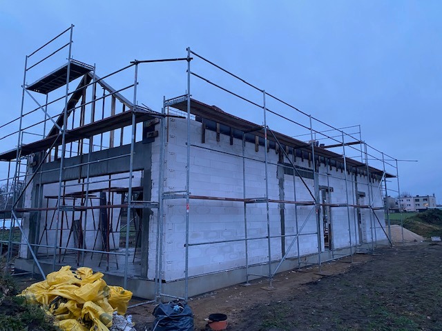 Budowa domu z betonu komórkowego w Ostrowie Wielkopolskim, widoczne rusztowania i konstrukcja dachu w trakcie montażu, niebieskie niebo w tle.