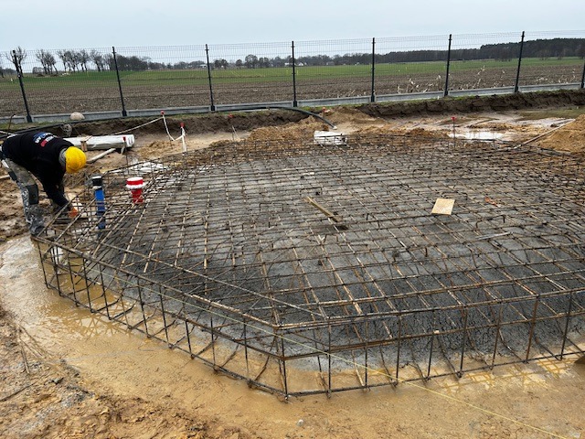 Pracownik w żółtym kasku przy zbrojeniu okrągłego fundamentu na błotnistym terenie budowy, widoczne stalowe pręty i częściowo zalany beton.