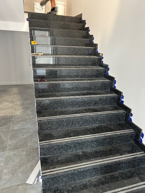 Granitowe schody z antypoślizgowymi paskami podczas montażu, widoczne niebieskie kliny dystansowe i narzędzia murarskie na stopniach.