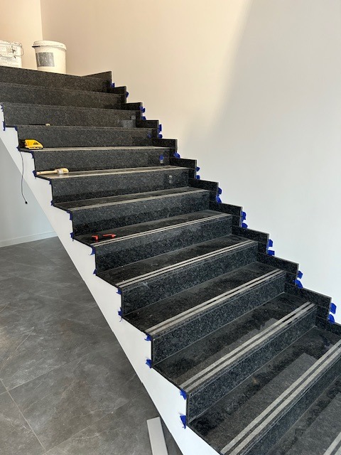 Schody wewnętrzne wyłożone ciemnym granitem z jasnymi paskami antypoślizgowymi, zabezpieczone niebieską taśmą malarską na krawędziach stopni, widoczne narzędzia i pojemnik z farbą wskazują...
