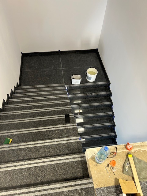 Schody wyłożone ciemnym gresem z białymi paskami antypoślizgowymi, widoczne narzędzia i materiały budowlane, etap prac wykończeniowych.