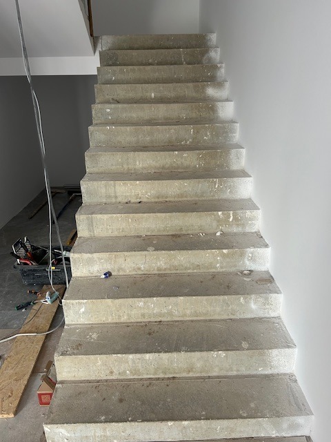 Surowe betonowe schody wewnętrzne w trakcie budowy, widoczne narzędzia i materiały budowlane, szare ściany w tle, robocze oświetlenie.