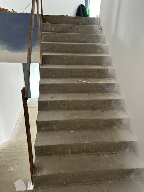Surowe betonowe schody w trakcie budowy, z prowizoryjną metalową barierką i fragmentem pomalowanej ściany w tle.