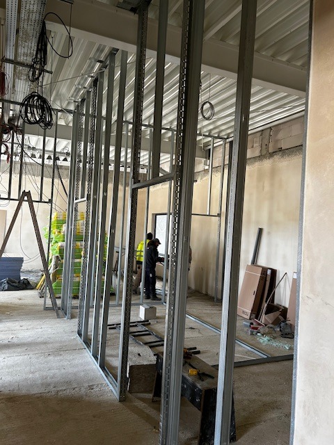 Wnętrze w trakcie tworzenia ścian działowych z metalowych profili, widoczni pracownicy w odblaskowych kamizelkach, surowy stan wykończenia, widoczne instalacje pod sufitem.