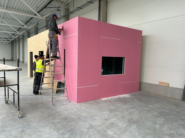 Pracownik na drabinie montuje różowe płyty gipsowo-kartonowe z oknem wewnątrz hali, drugi pracownik w żółtej kamizelce obserwuje, w tle konstrukcja z płyt OSB.
