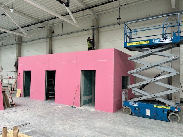 Wnętrze hali z budową ścian działowych z różowych płyt gipsowo-kartonowych, widoczny podnośnik nożycowy i pracownicy na rusztowaniu.