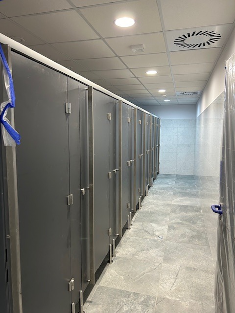 Rząd szarych kabin sanitarnych z metalowymi uchwytami w nowocześnie wykończonej toalecie publicznej, widok z perspektywy korytarza, podłoga wyłożona płytkami imitującymi kamień.