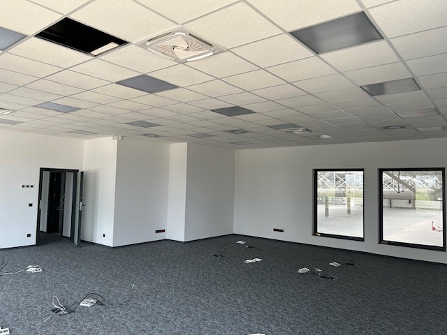 Pusta przestrzeń biurowa z szarą wykładziną, podwieszanym sufitem z panelami, otwartymi drzwiami i widokiem z okna na zewnątrz, przygotowana do prac wykończeniowych.