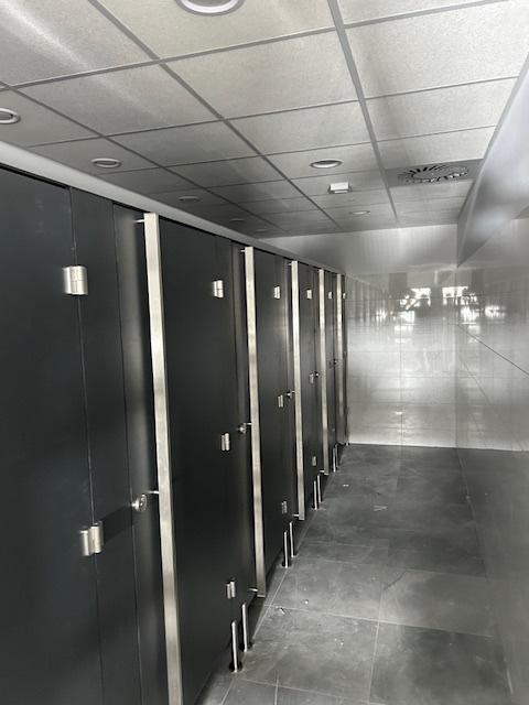 Rząd kabin toaletowych z szarymi drzwiami i metalowymi okuciami, widok wzdłuż długiego korytarza z szarą podłogą i sufitem kasetonowym.