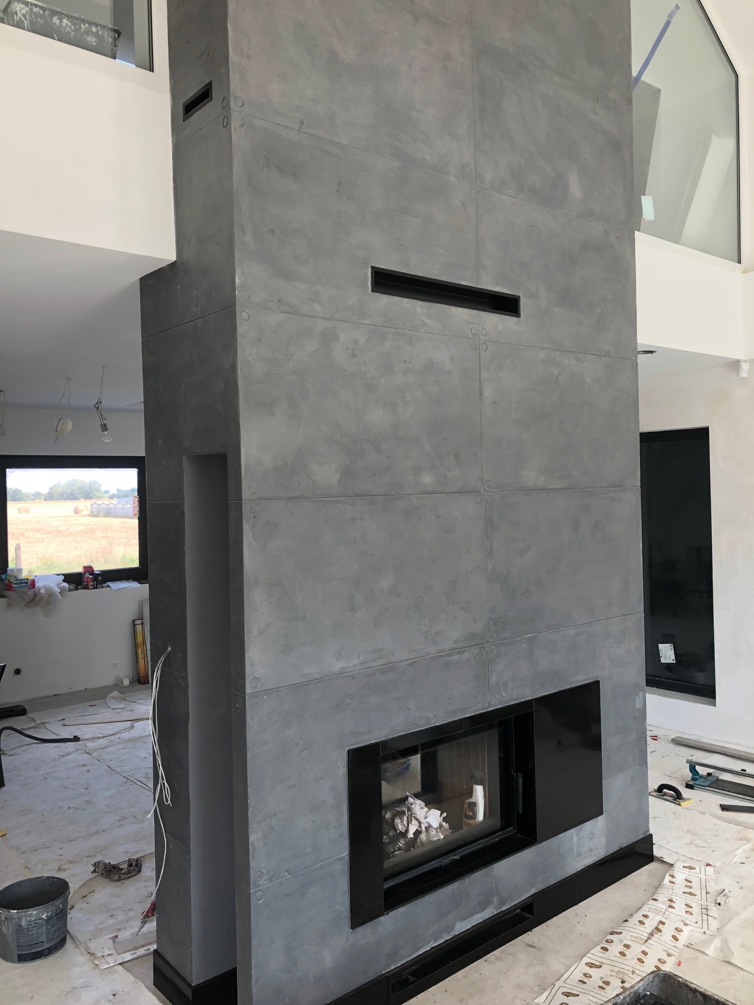 Wysoki kominek z szarych płyt imitujących beton, z czarnym wkładem kominkowym i czarnymi wstawkami, w surowym wnętrzu w trakcie prac wykończeniowych.