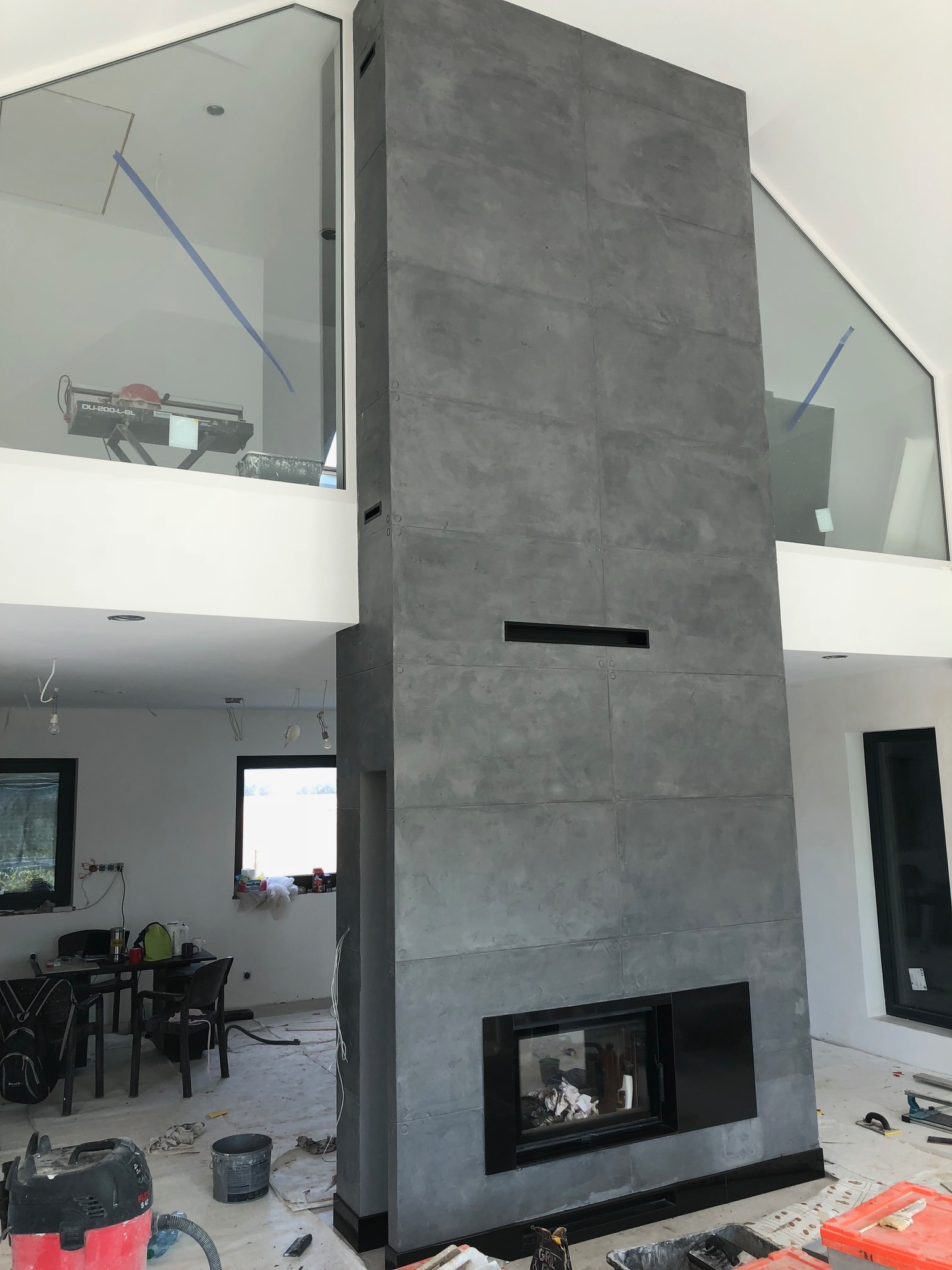 Wysoki komin obłożony szarymi płytami imitującymi beton w surowym wnętrzu domu z dużymi oknami, widoczne narzędzia i materiały budowlane.