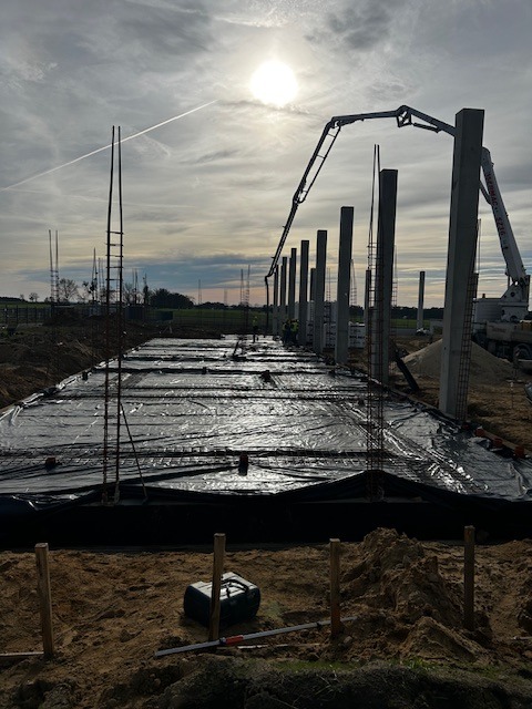 Wylewanie fundamentów pod budynek z betonowymi słupami, widoczny dźwig z pompą do betonu na tle zachmurzonego nieba z przebijającym się słońcem.