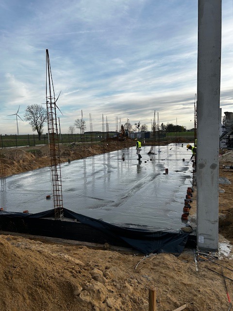 Wylewanie fundamentów pod budynek z widocznymi zbrojeniami, pracownikami w odblaskowych kamizelkach i turbinami wiatrowymi w oddali, pochmurne niebo.