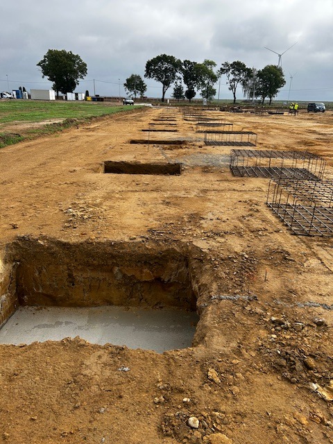 Wykopy pod fundamenty z częściowo wylanym betonem, widoczne zbrojenia i turbina wiatrowa w tle.