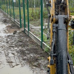 KAMIL ZAWISZA - Prace ziemne z użyciem minikoparki podczas budowy ogrodzenia z zielonych słupków i prefabrykowanej podmurówki w błotnistym terenie.