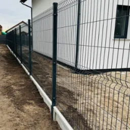 Nowoczesne ogrodzenie panelowe w kolorze grafitowym, zamontowane na betonowej podmurówce, otaczające posesję z białym budynkiem w tle.