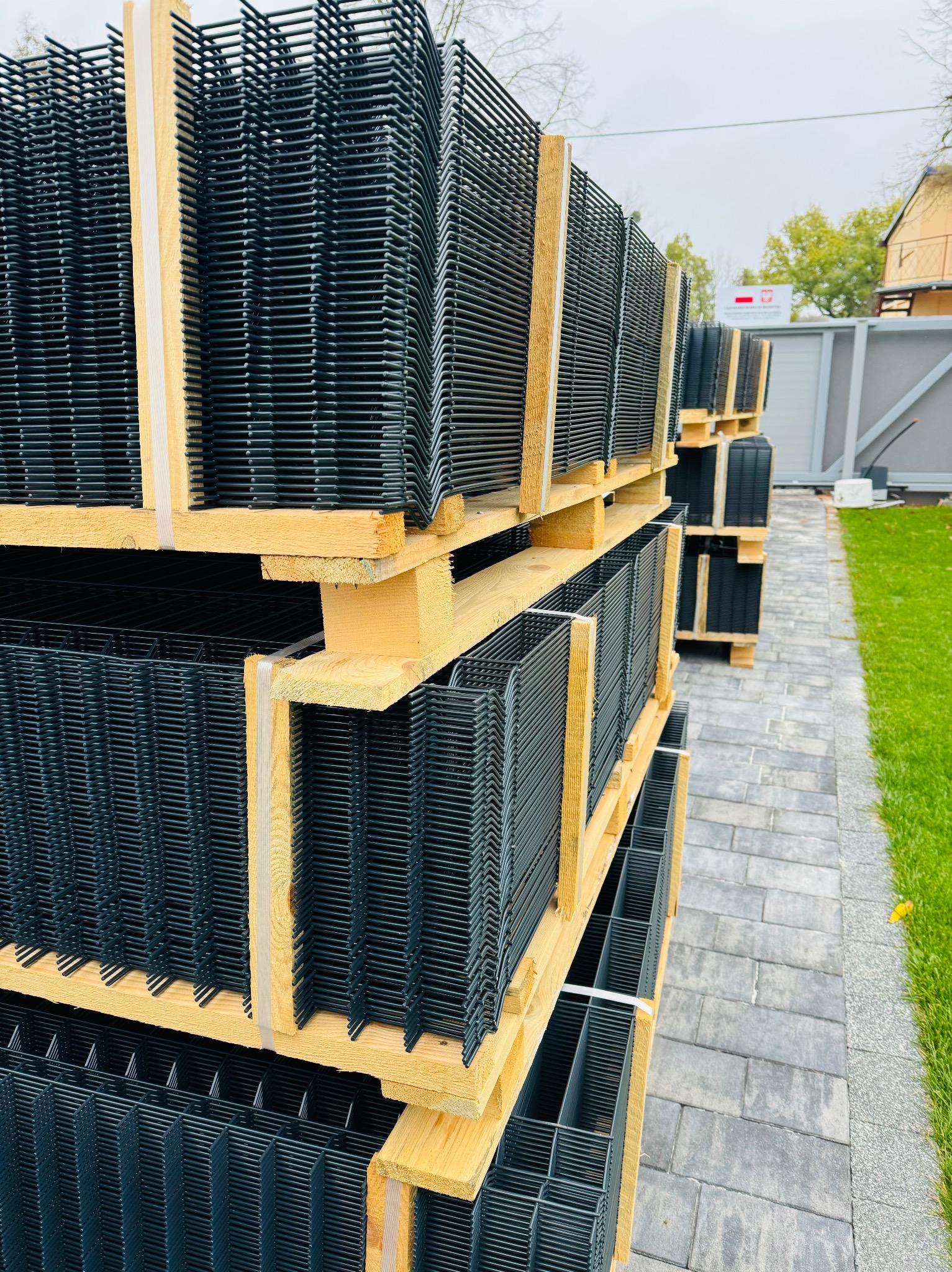 Stosy paneli ogrodzeniowych zgrzewanych z drutu, ułożone na drewnianych paletach, czekające na montaż, widoczne na tle kostki brukowej i trawnika.