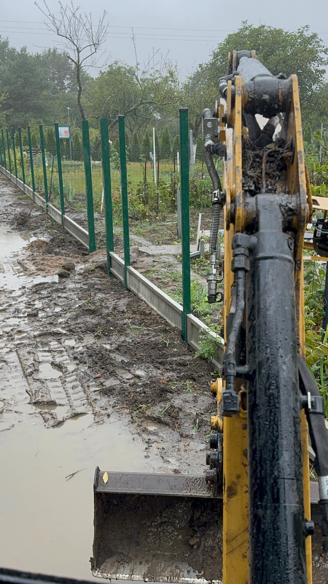 Prace ziemne z użyciem minikoparki podczas budowy ogrodzenia z zielonych słupków i prefabrykowanej podmurówki w błotnistym terenie.