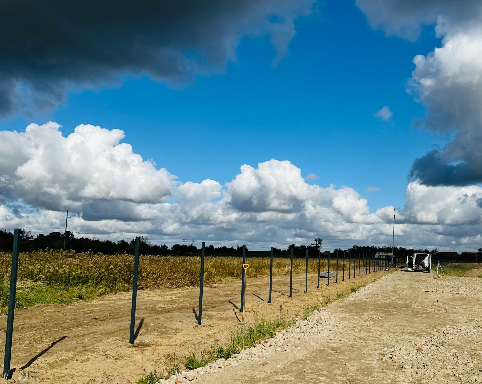 Wkopane słupki pod ogrodzenie na tle pola i malowniczego nieba z chmurami, widoczny samochód dostawczy i narzędzia.