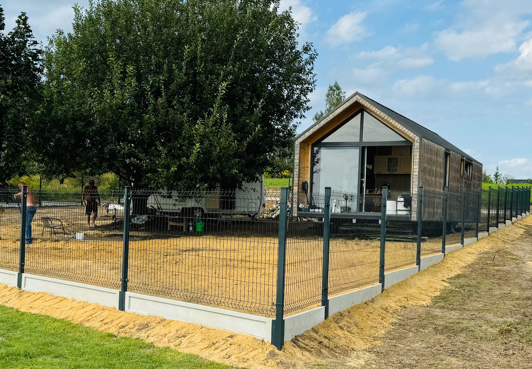 Nowo zainstalowane, ciemne ogrodzenie panelowe z podmurówką betonową otacza nowoczesny, drewniany dom z dużymi oknami, widoczny trawnik i drzewa w tle, dwóch mężczyzn w oddali.