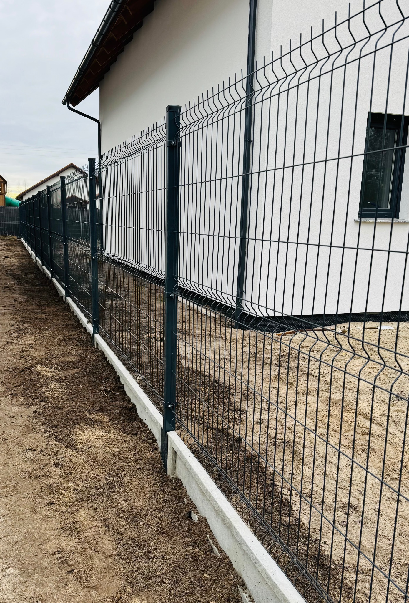 Nowoczesne ogrodzenie panelowe w kolorze grafitowym, zamontowane na betonowej podmurówce, otaczające posesję z białym budynkiem w tle.