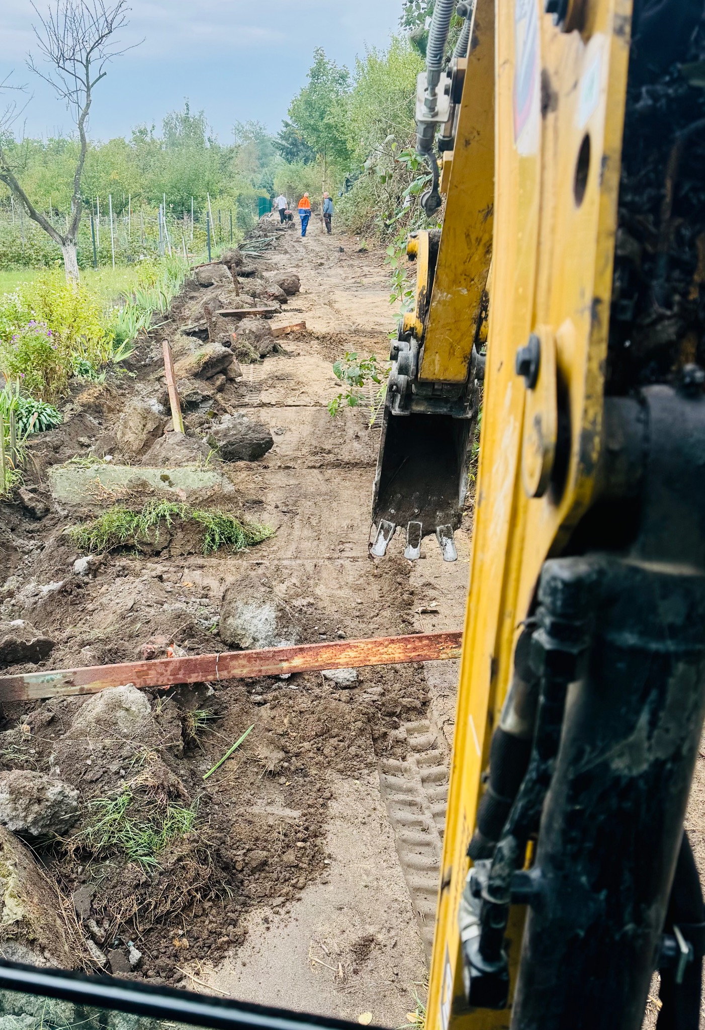 Żółta koparka w trakcie prac ziemnych przy budowie ogrodzenia, widoczne wyrównane podłoże, kamienie i fragmenty drewnianych elementów, w tle pracownicy i siatka ogrodzeniowa.