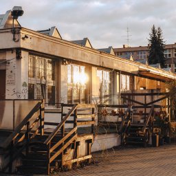 In Boiling Water Company - Dwukondygnacyjny budynek z oknami w metalowych kratach, oświetlony zachodzącym słońcem, z drewnianymi schodami wejściowymi i graffiti na ścianie.