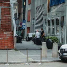 In Boiling Water Company - Ulica miejska z budynkami biurowymi, zaparkowanymi samochodami, schodami z metalową konstrukcją i dwoma mężczyznami rozmawiającymi na chodniku.