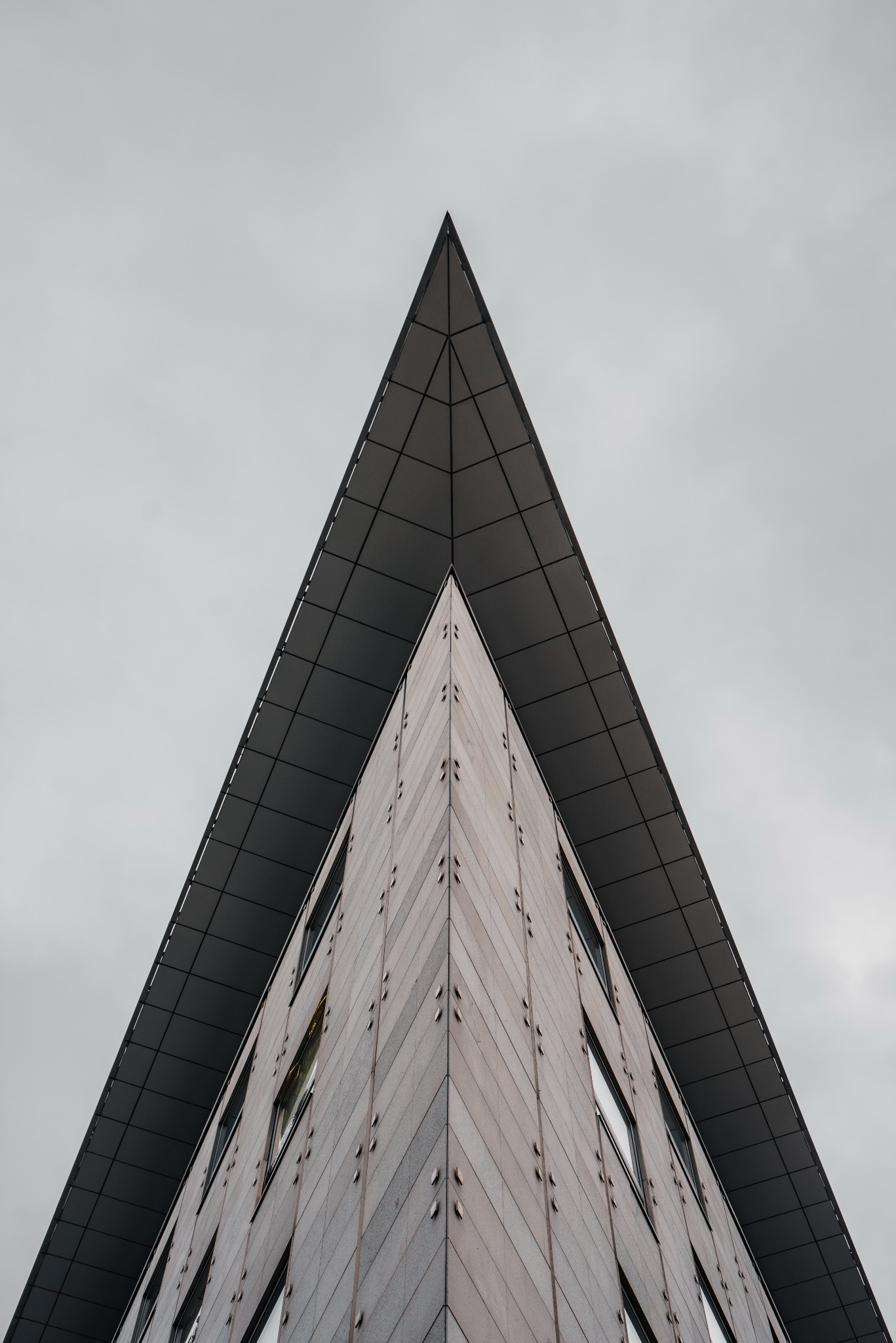 Szczyt nowoczesnego budynku biurowego w Warszawie, widziany z dołu pod ostrym kątem, z wyraźną trójkątną strukturą dachu i elewacją wykończoną w odcieniach szarości i beżu. Widoczne regularne...