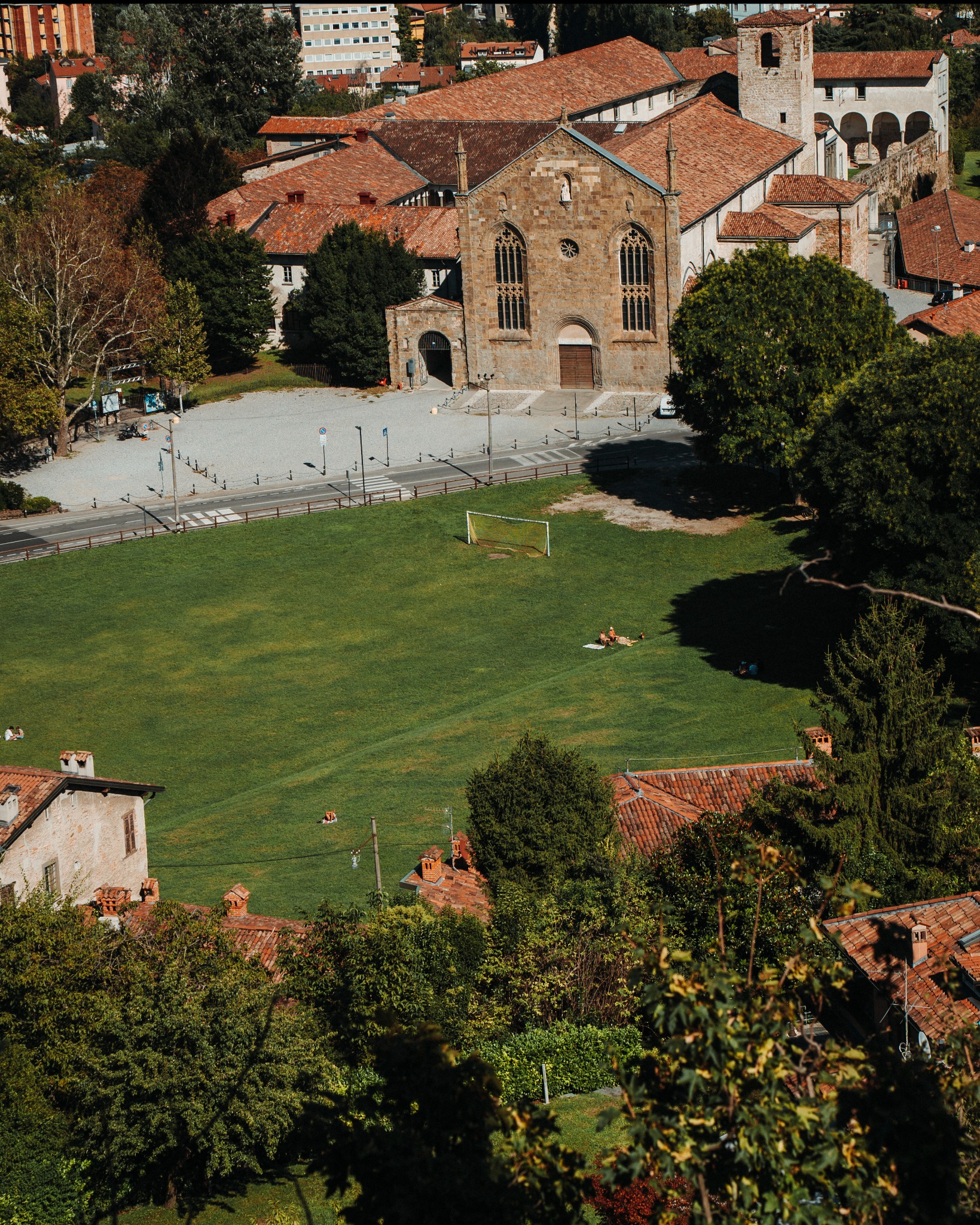 Widok z góry na włoskie miasto Bergamo, z zielonym trawnikiem na pierwszym planie, średniowieczną architekturą z czerwonymi dachami w tle oraz osobami odpoczywającymi na trawie w słoneczny dzień.