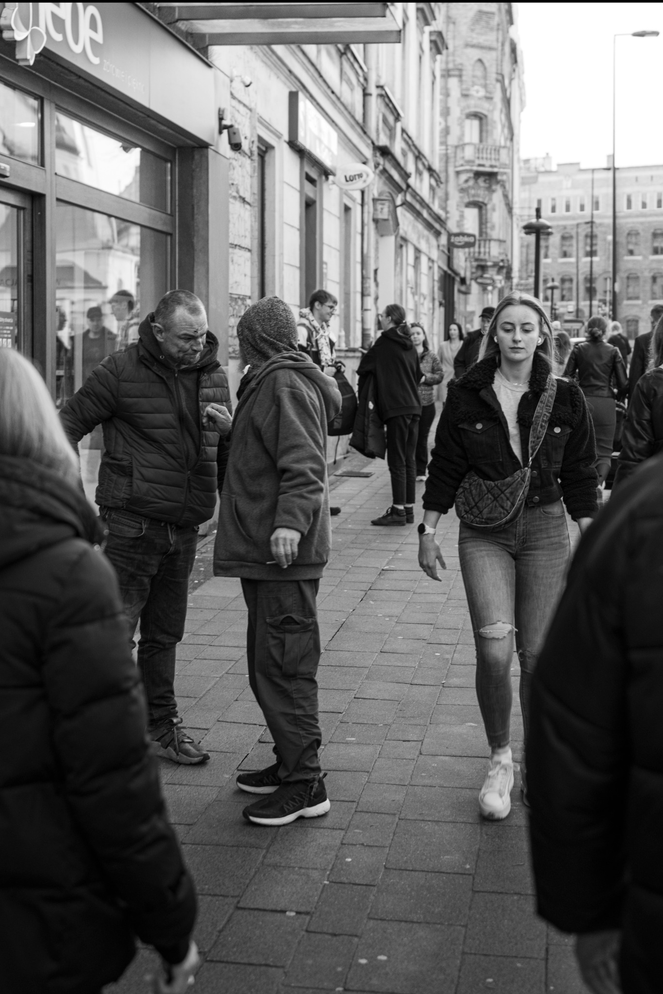 Czarno-białe zdjęcie uliczne przedstawiające młodą kobietę w dżinsach z dziurami i kurtce z barankiem, idącą chodnikiem w otoczeniu innych przechodniów i budynków w tle.