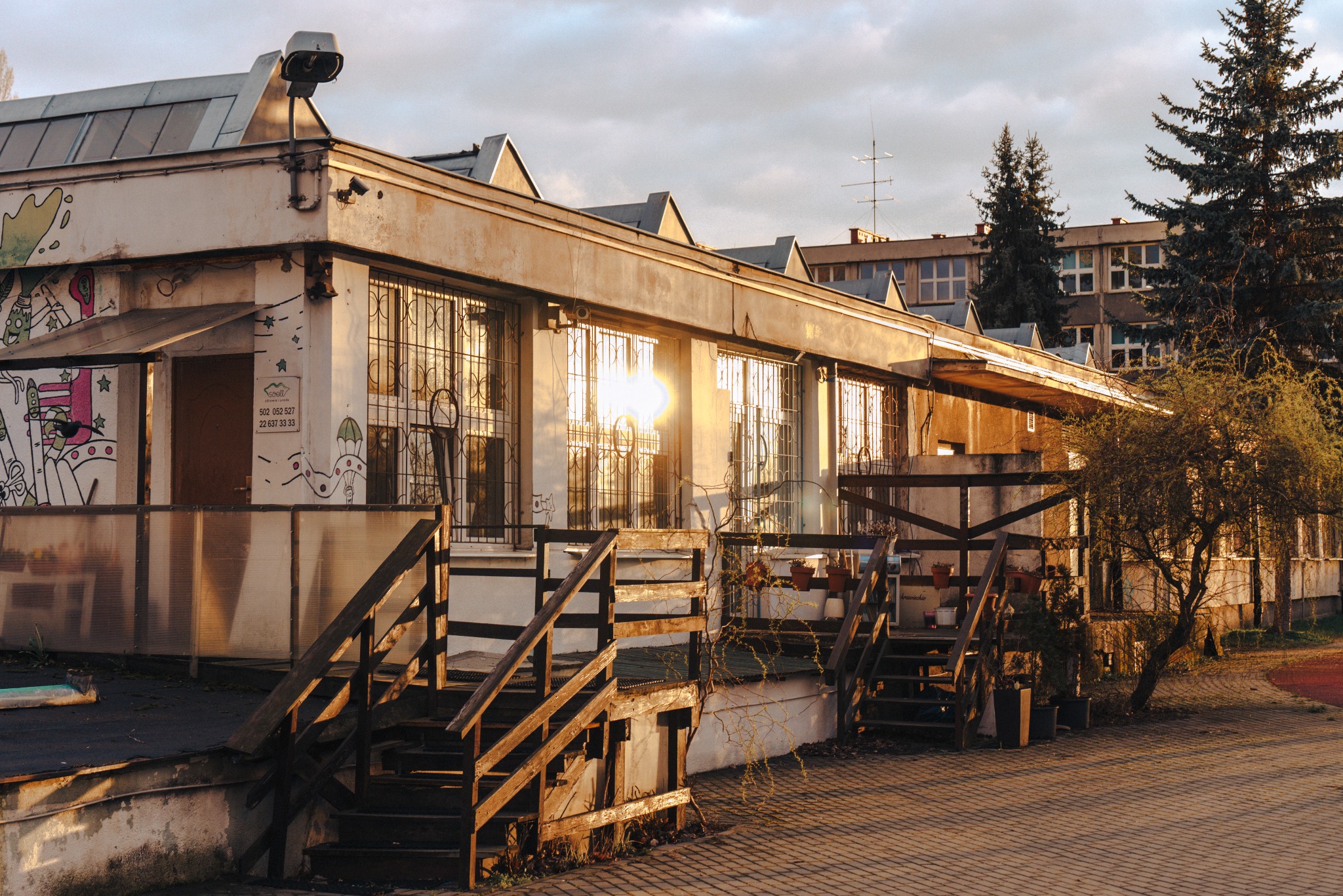 Dwukondygnacyjny budynek z oknami w metalowych kratach, oświetlony zachodzącym słońcem, z drewnianymi schodami wejściowymi i graffiti na ścianie.