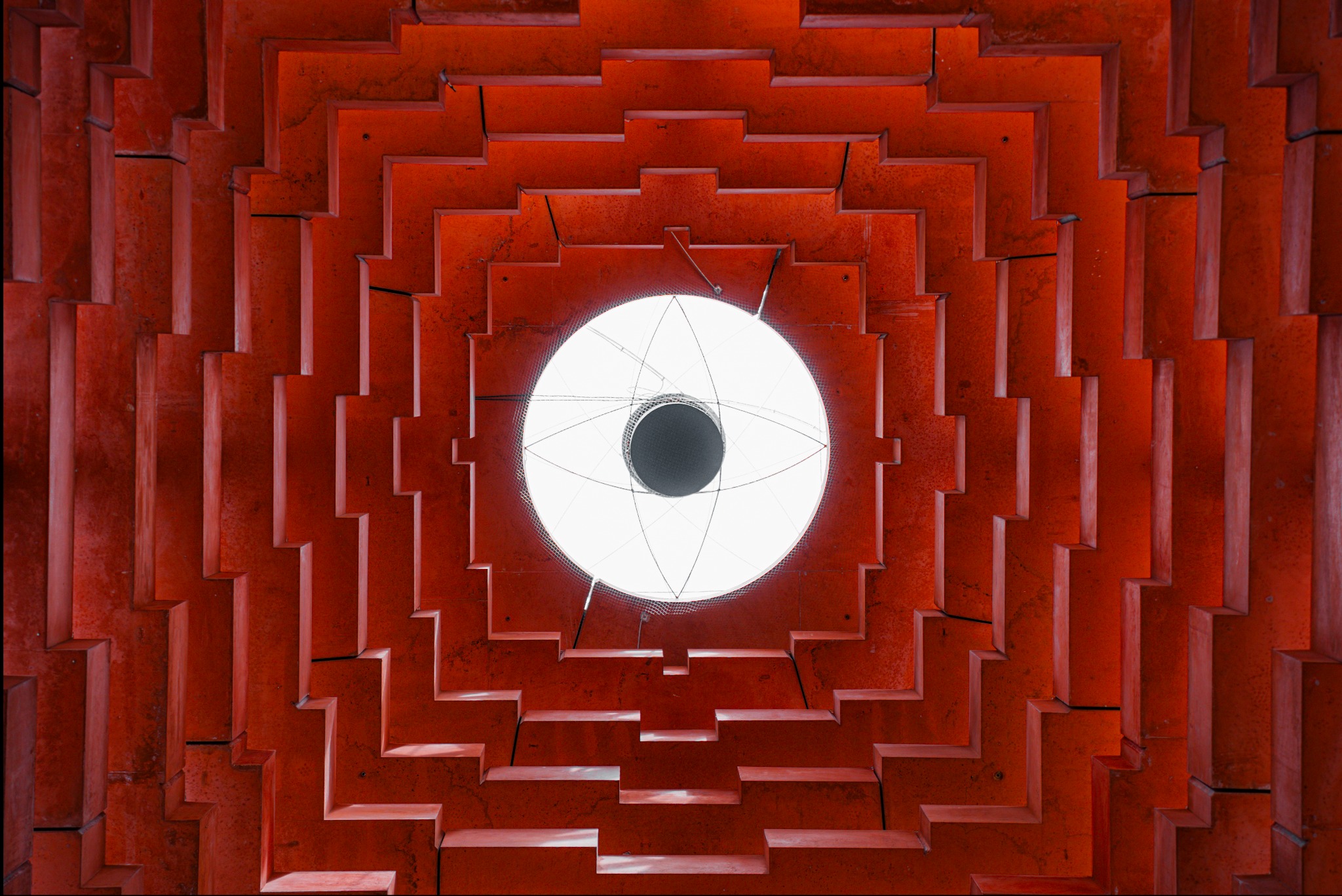 Widok z dołu na nowoczesną, czerwoną konstrukcję sufitową z centralnie umieszczonym okrągłym świetlikiem, otoczonym geometrycznym wzorem schodkowych elementów.