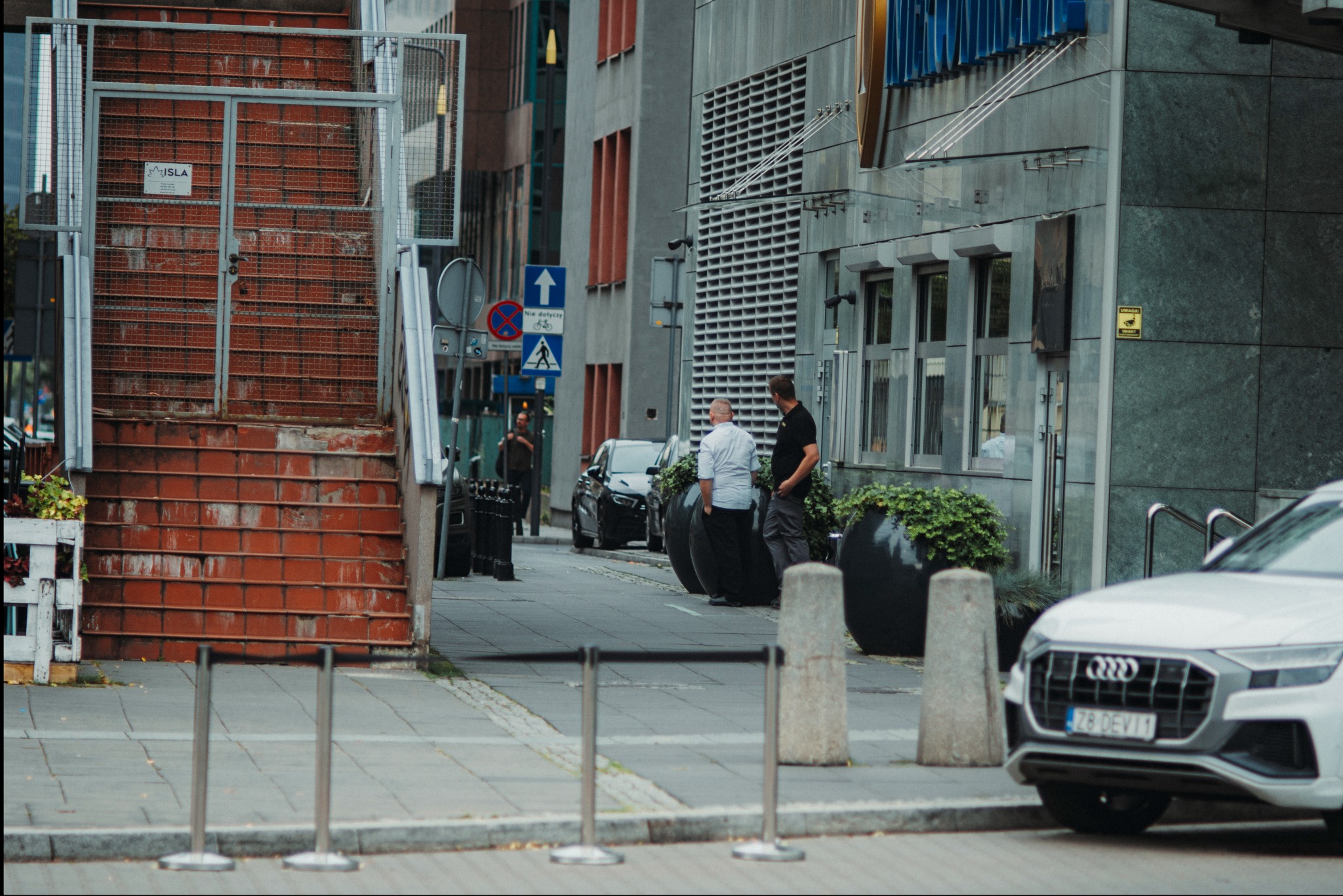 Ulica miejska z budynkami biurowymi, zaparkowanymi samochodami, schodami z metalową konstrukcją i dwoma mężczyznami rozmawiającymi na chodniku.