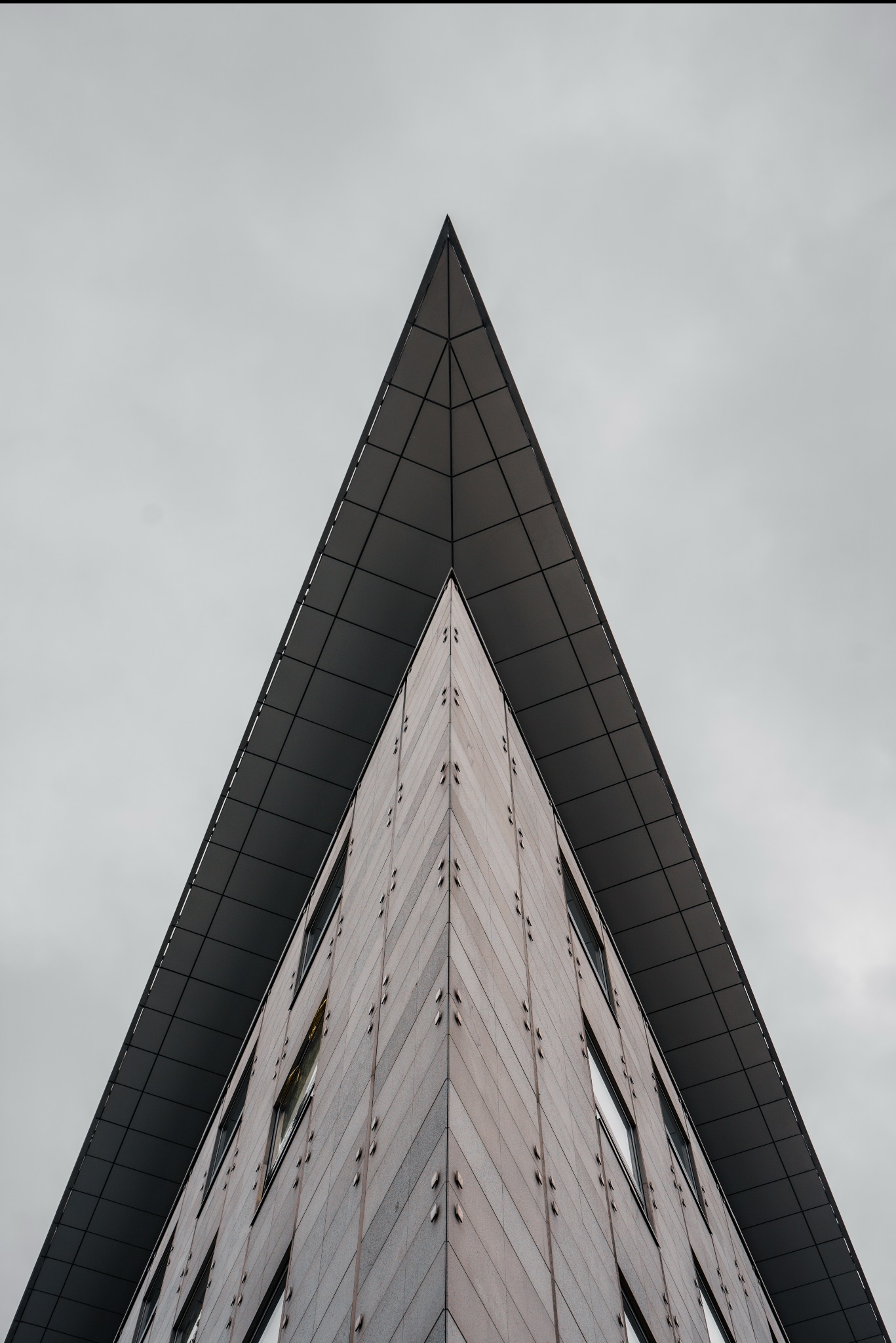 Nietypowe ujęcie wertykalne na szczyt budynku o trójkątnym zwieńczeniu, z widocznymi oknami i elewacją w odcieniach szarości, na tle pochmurnego nieba.