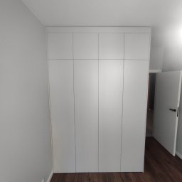 Michał PIĘTKA - Biała szafa wnękowa z czterema drzwiami, zamontowana od podłogi do sufitu w pomieszczeniu z brązową podłogą i szarą ścianą, z otwartymi białymi drzwiami w tle.