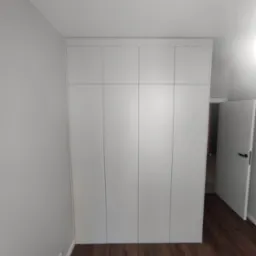 Biała szafa wnękowa z czterema drzwiami, zamontowana od podłogi do sufitu w pomieszczeniu z brązową podłogą i szarą ścianą, z otwartymi białymi drzwiami w tle.