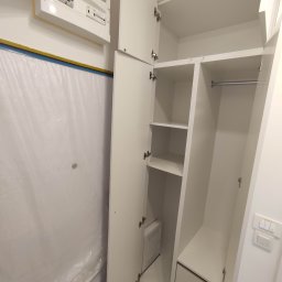 Michał PIĘTKA - Wnętrze białej szafy z półkami i szufladami, widoczny panel elektryczny na ścianie zabezpieczonej folią malarską.