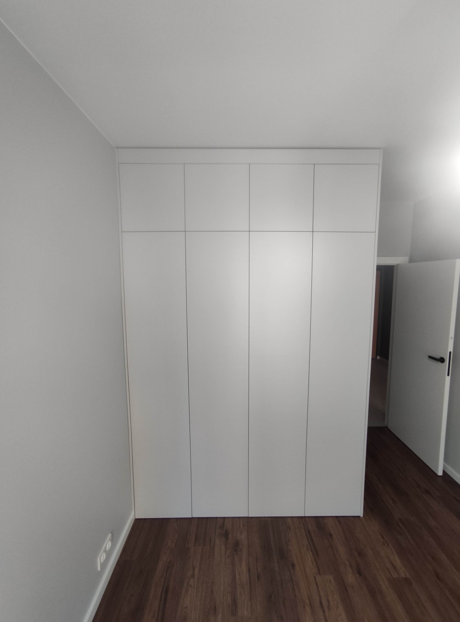 Biała szafa wnękowa z czterema drzwiami, zamontowana od podłogi do sufitu w pomieszczeniu z brązową podłogą i szarą ścianą, z otwartymi białymi drzwiami w tle.