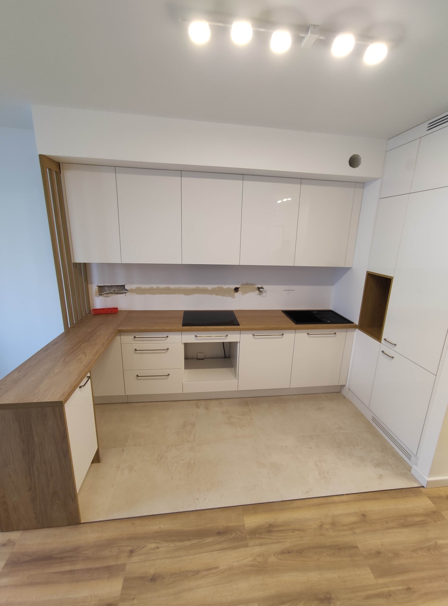 Wnętrze kuchni w trakcie montażu, widoczne białe, lakierowane szafki kuchenne z drewnianym blatem, płyty indukcyjne i niezamaskowane otwory w ścianie nad blatem.