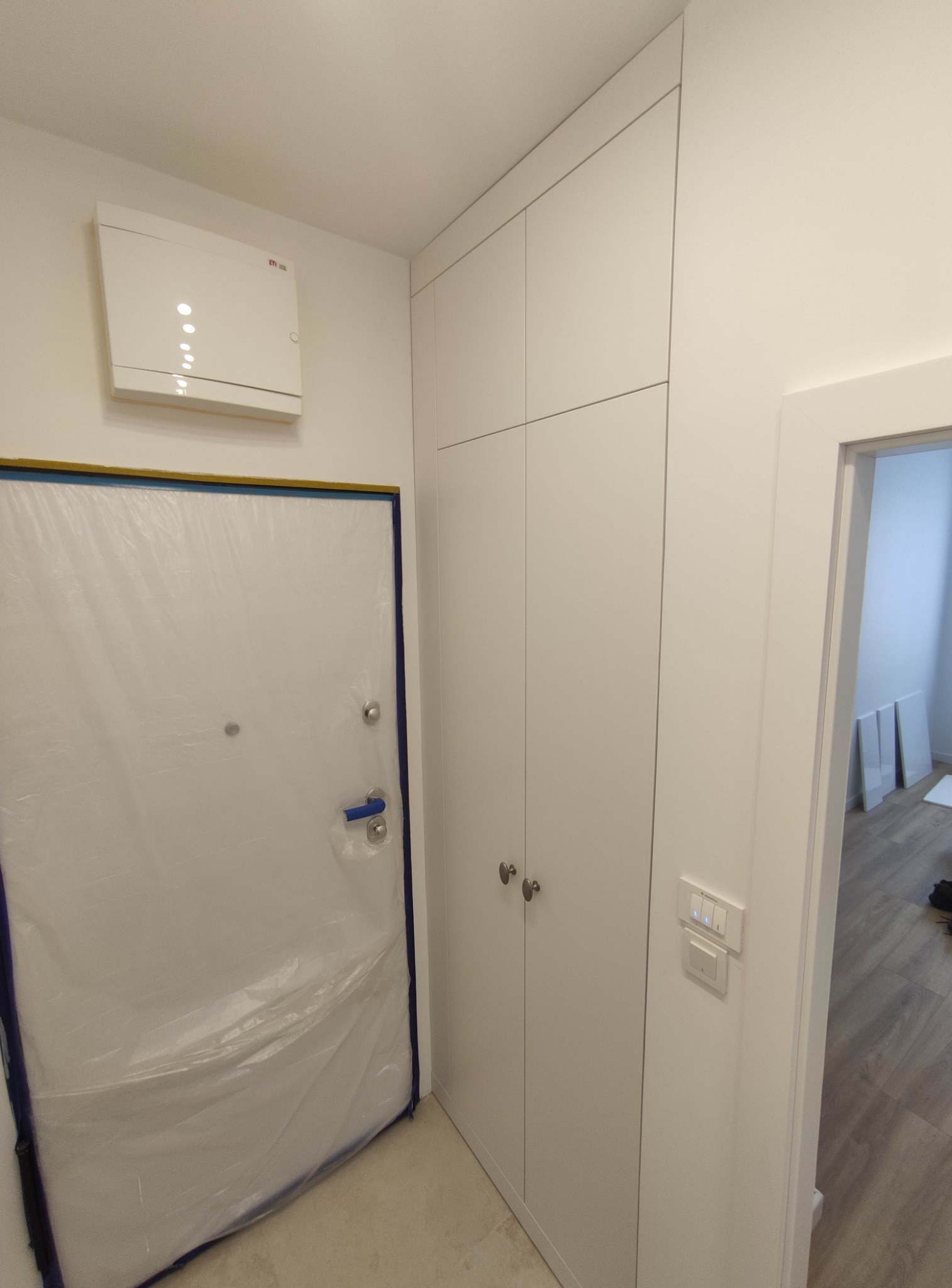Biała, wysoka szafa w przedpokoju, z widocznym zabezpieczeniem drzwi folią malarską, obok włączniki światła, w tle widoczne elementy podłogi i drzwi.