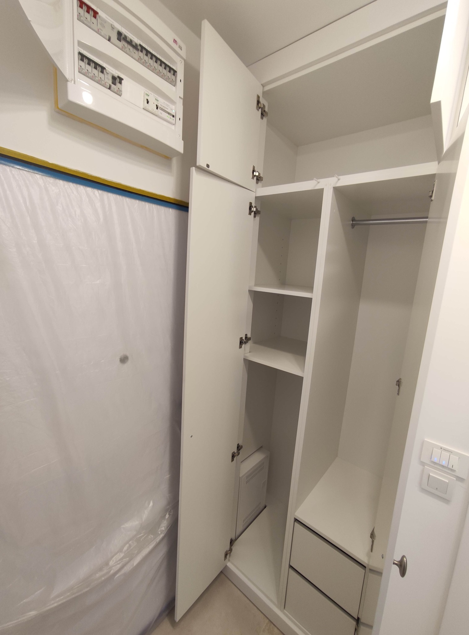 Wnętrze białej szafy z półkami i szufladami, widoczny panel elektryczny na ścianie zabezpieczonej folią malarską.