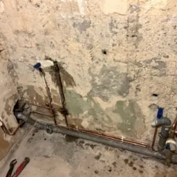 Instalacja wodna z rur miedzianych i szarych rur PCV w surowym wnętrzu ze ścianami z odpadającym tynkiem, widoczne narzędzia hydrauliczne