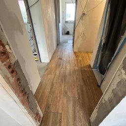 SKY HOME Wykończenia Wnętrz Dominik Lipa - Korytarz w trakcie remontu: ułożona drewnopodobna podłoga z płytek, ściany w stanie surowym, widoczne kable i elementy konstrukcyjne. Perspektywa z wnętrza pomieszczenia.