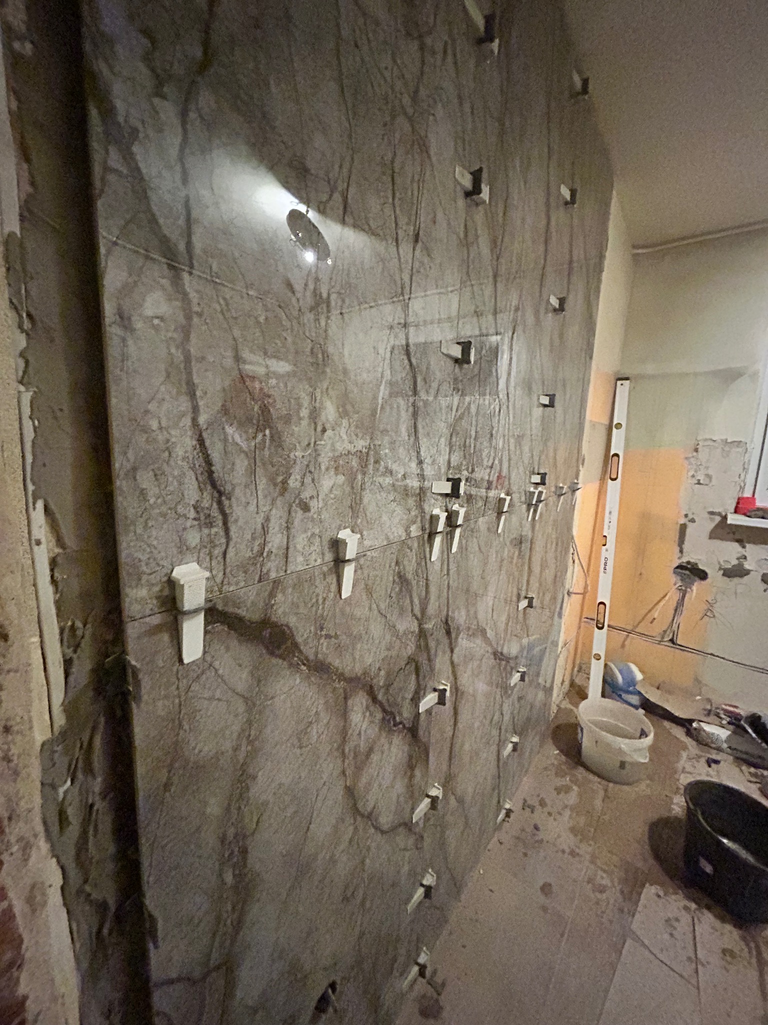 Montaż płytek imitujących marmur na ścianie łazienki z użyciem klipsów poziomujących, widoczny poziomica i akcesoria glazurnicze w tle.