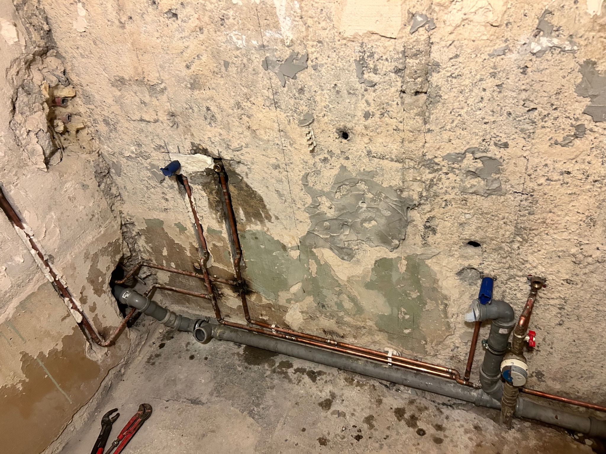 Instalacja wodna z rur miedzianych i szarych rur PCV w surowym wnętrzu ze ścianami z odpadającym tynkiem, widoczne narzędzia hydrauliczne